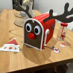 May include: A red and brown mailbox shaped like a reindeer with large eyes, a red nose, and antlers. A Santa Claus cutout, a snowman cutout, and a bottle with a Santa Claus graphic are also on the wooden table.