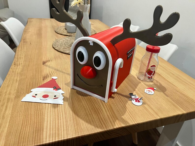 May include: A red and brown mailbox shaped like a reindeer with large eyes, a red nose, and antlers. A Santa Claus cutout, a snowman cutout, and a bottle with a Santa Claus graphic are also on the wooden table.