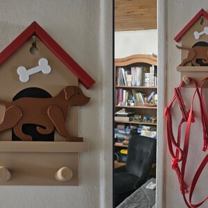 May include: Wooden dog-themed wall-mounted leash holder. The holder features a brown dog silhouette, a white bone, and a red roof. Two wooden pegs are below for hanging leashes. A red leash hangs on a similar holder.