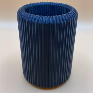 May include: A cylindrical, navy blue container with vertical ribbed detailing. The object is made of a solid material and has a smooth, matte finish. The container is suitable for holding pencils, pens, or small items.