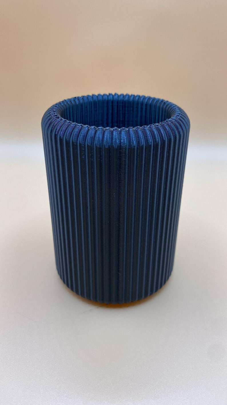 May include: A cylindrical, navy blue container with vertical ribbed detailing. The object is made of a solid material and has a smooth, matte finish. The container is suitable for holding pencils, pens, or small items.