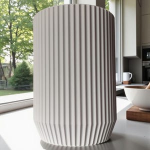 May include: A tall, cylindrical, off-white vase with vertical ribbed detailing. The vase has a geometric base and stands on a light-colored surface. The background includes a window with a view of trees and a kitchen setting.