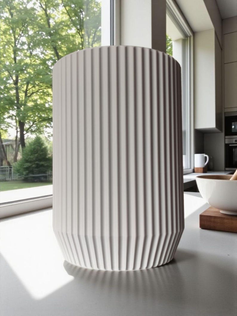 May include: A tall, cylindrical, off-white vase with vertical ribbed detailing. The vase has a geometric base and stands on a light-colored surface. The background includes a window with a view of trees and a kitchen setting.