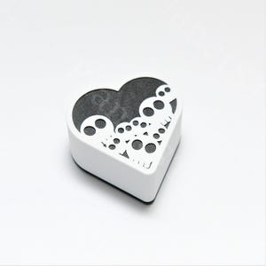 May include: A white, heart-shaped box with a black interior. The top of the box features a decorative cut-out design of skulls. The box is sitting on a white surface.