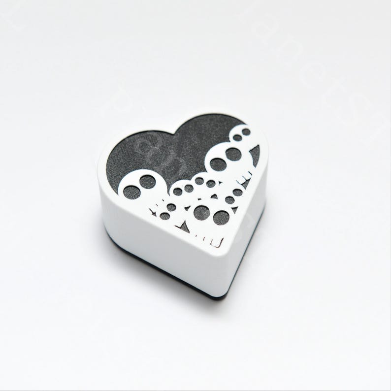 May include: A white, heart-shaped box with a black interior. The top of the box features a decorative cut-out design of skulls. The box is sitting on a white surface.