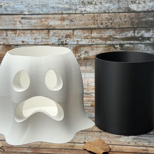 May include: A white ghost-shaped Halloween decoration with oval eye cutouts and a curved mouth. A black cylindrical container sits beside it. The background features a wooden surface and pumpkins.