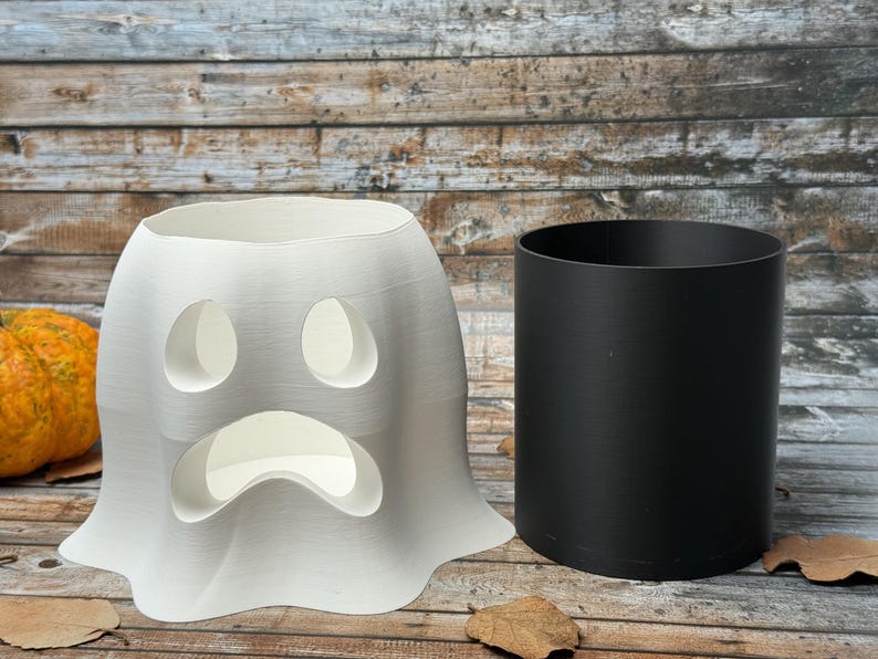 May include: A white ghost-shaped Halloween decoration with oval eye cutouts and a curved mouth. A black cylindrical container sits beside it. The background features a wooden surface and pumpkins.