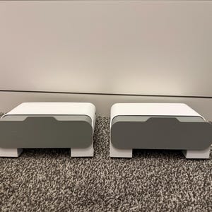 May include: Two matching white and gray storage boxes with a modern design. Each box has a rectangular shape with a recessed gray front panel and sits on a white base. The boxes are on a patterned carpet.