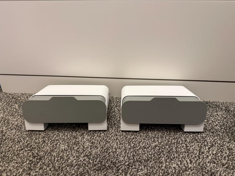 May include: Two matching white and gray storage boxes with a modern design. Each box has a rectangular shape with a recessed gray front panel and sits on a white base. The boxes are on a patterned carpet.