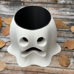 May include: A white ghost-shaped planter with a black interior. The planter has two circular eye holes and a curved mouth. An orange pumpkin and dried leaves are in the background. This Halloween-themed planter is perfect for seasonal decor.