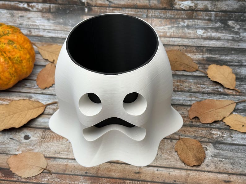 May include: A white ghost-shaped planter with a black interior. The planter has two circular eye holes and a curved mouth. An orange pumpkin and dried leaves are in the background. This Halloween-themed planter is perfect for seasonal decor.