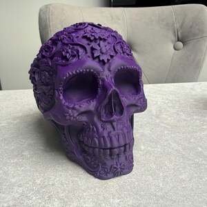 May include: A decorative purple skull with floral and scrollwork details. The skull has large eye sockets, a prominent nose cavity, and a detailed jawline. The skull is sitting on a light gray surface.