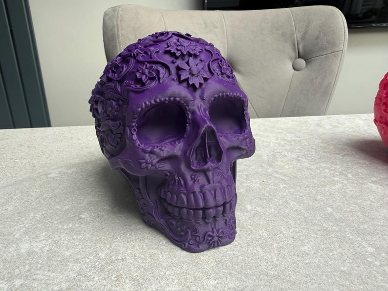 May include: A decorative purple skull with floral and scrollwork details. The skull has large eye sockets, a prominent nose cavity, and a detailed jawline. The skull is sitting on a light gray surface.