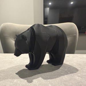 May include: A black bear figurine with a geometric design. The bear is standing on all fours, with its head slightly lowered. The figurine is made of a solid black material and casts a shadow on the surface below.