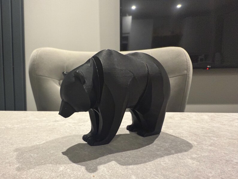 May include: A black bear figurine with a geometric design. The bear is standing on all fours, with its head slightly lowered. The figurine is made of a solid black material and casts a shadow on the surface below.