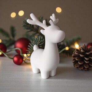 May include: A white ceramic reindeer figurine with antlers, a rounded body, and four legs. The reindeer is a Christmas decoration, with red ornaments, pine needles, and a pine cone in the background. Warm lights are strung around the scene.