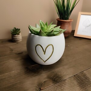 Op de afbeelding: Een witte, ronde keramische plantenbak met een gouden hartontwerp, met een groene vetplant. Extra planten in terracotta potten en een ingelijste bladtekening zijn zichtbaar op de achtergrond. De plantenbak staat op een houten oppervlak.