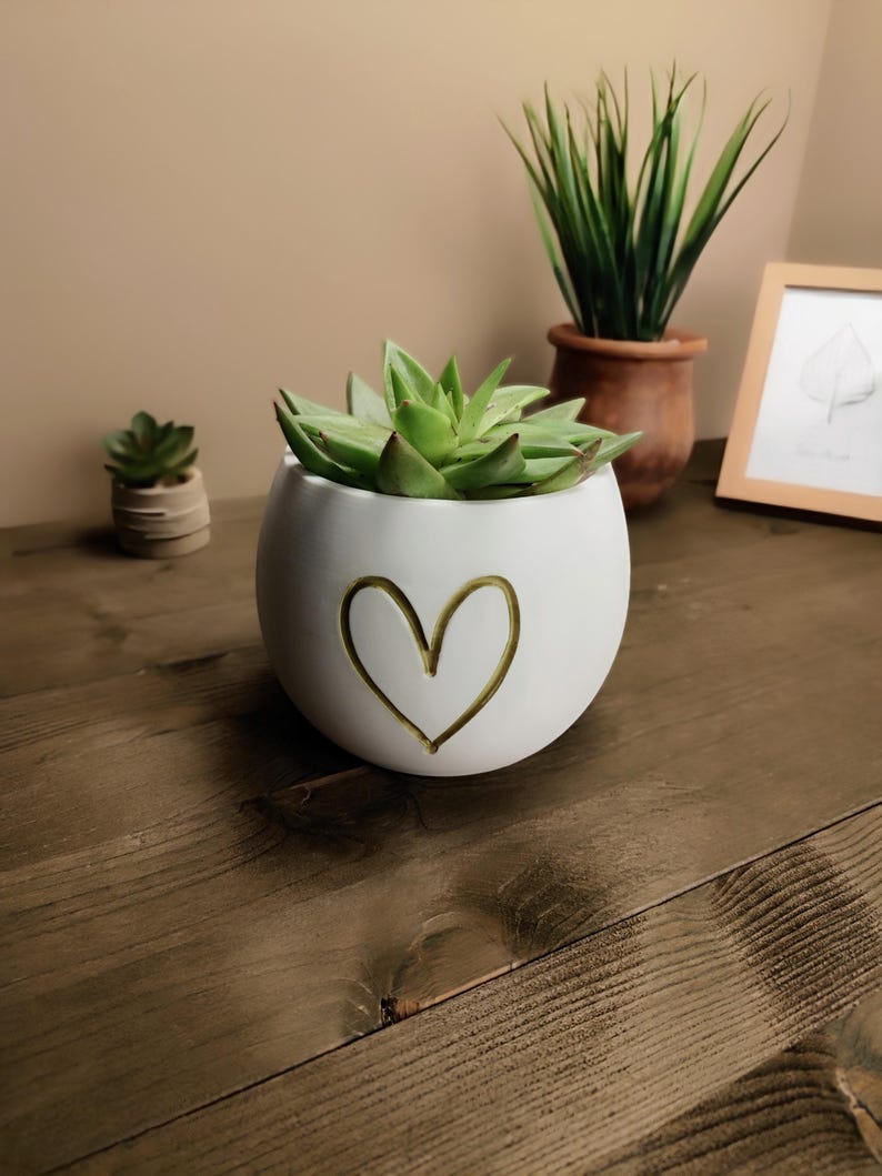 Op de afbeelding: Een witte, ronde keramische plantenbak met een gouden hartontwerp, met een groene vetplant. Extra planten in terracotta potten en een ingelijste bladtekening zijn zichtbaar op de achtergrond. De plantenbak staat op een houten oppervlak.