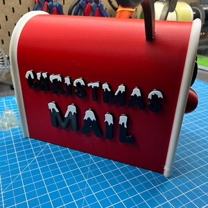 May include: A red Christmas mailbox with white trim and the words "CHRISTMAS MAIL" in black letters with white snow accents. The mailbox has a red knob and a black handle.