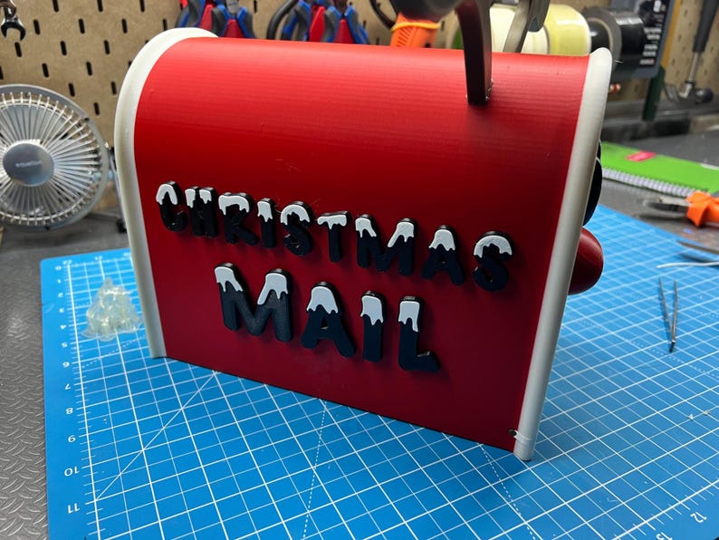 May include: A red Christmas mailbox with white trim and the words "CHRISTMAS MAIL" in black letters with white snow accents. The mailbox has a red knob and a black handle.