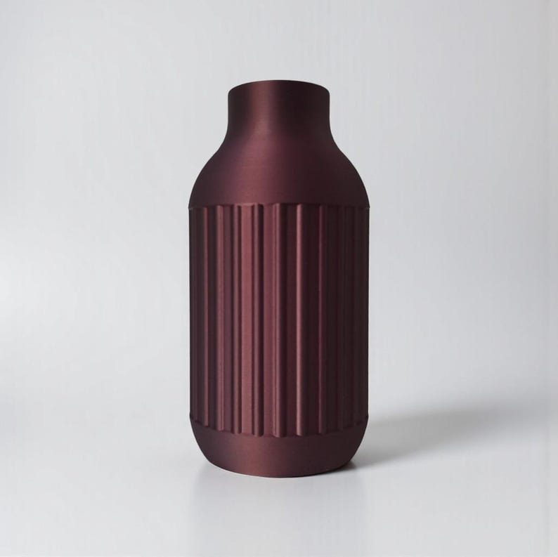 May include: A maroon-colored vase with a cylindrical body and a narrow neck. The vase features vertical, ridged detailing around the body, adding texture. The design is modern and minimalist, suitable for home decor.