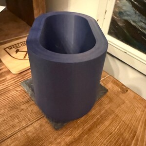 May include: A cylindrical, dark blue object with an oval opening, resting on a square, gray base. The object is made of a solid material and sits on a wooden surface. The interior of the object is also dark blue.