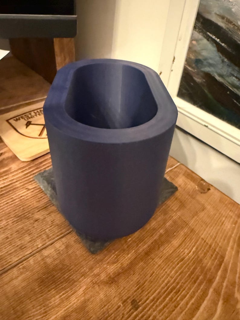 May include: A cylindrical, dark blue object with an oval opening, resting on a square, gray base. The object is made of a solid material and sits on a wooden surface. The interior of the object is also dark blue.
