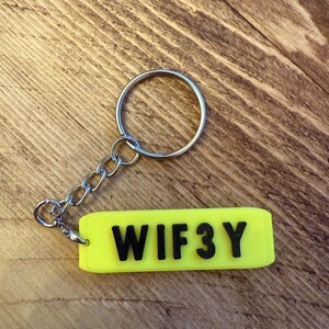 May include: A neon yellow keychain tag with the word "WIF3Y" in black block letters. The tag is attached to a silver chain and keyring. The background is a wooden surface.