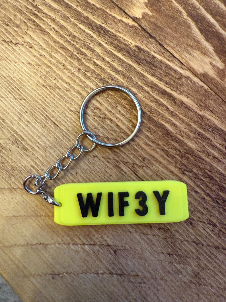 May include: A neon yellow keychain tag with the word "WIF3Y" in black block letters. The tag is attached to a silver chain and keyring. The background is a wooden surface.
