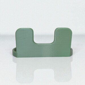 May include: A sage green, U-shaped desk organizer. The organizer has a rectangular base and two upright sections connected by a horizontal space. The item is made of a solid material and sits on a reflective surface.
