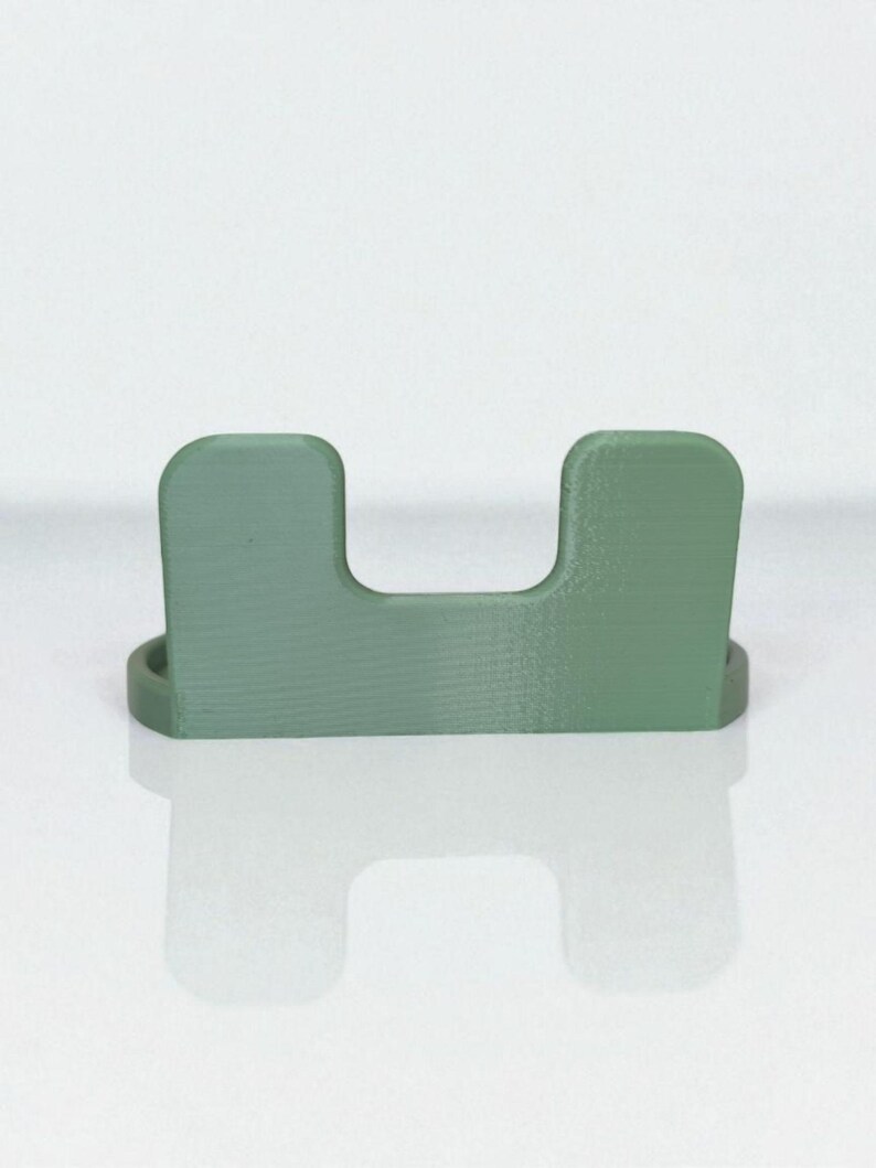May include: A sage green, U-shaped desk organizer. The organizer has a rectangular base and two upright sections connected by a horizontal space. The item is made of a solid material and sits on a reflective surface.
