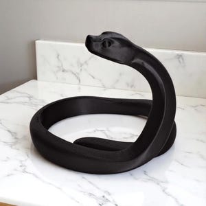 Puede incluir: Escultura de serpiente negra enroscada. La cabeza est&aacute; levantada. La escultura est&aacute; hecha de un material negro mate y descansa sobre una superficie de m&aacute;rmol blanco. La cabeza de la serpiente tiene detalles.