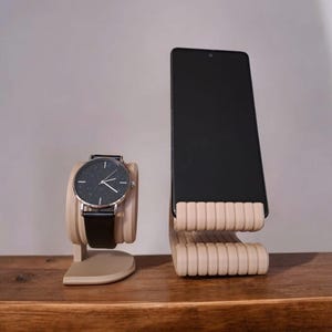 Puede incluir: Un reloj con esfera negra, correa de cuero negro y caja plateada se encuentra sobre un soporte de reloj beige. Un tel&eacute;fono inteligente negro descansa sobre un soporte de tel&eacute;fono beige con un dise&ntilde;o acanalado.