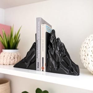 May include: Black mountain-shaped bookends holding two books on a white shelf. The bookends have a textured surface and are made of a solid material. The books are titled "THE STRENGTH IN OUR SCARS" and "KATHERINE MAY OBERWINTERN".