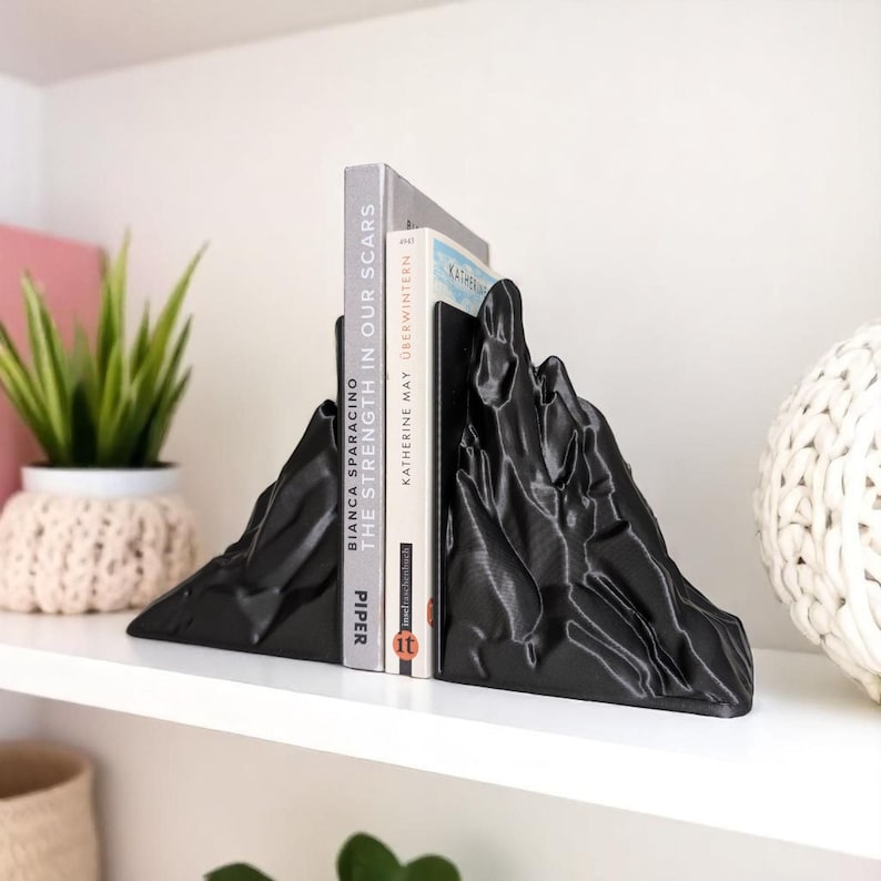 May include: Black mountain-shaped bookends holding two books on a white shelf. The bookends have a textured surface and are made of a solid material. The books are titled "THE STRENGTH IN OUR SCARS" and "KATHERINE MAY OBERWINTERN".