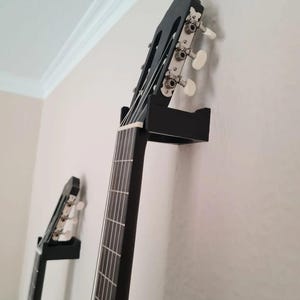 Op de afbeelding: Een zwarte akoestische gitaar hangt aan de muur met een zwarte gitaarhouder. De kop, stemknoppen, snaren en toets van de gitaar zijn zichtbaar. Een tweede gitaar is op de achtergrond gemonteerd.