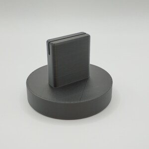 May include: A dark gray, 3D-printed phone stand. The stand has a rectangular slot to hold a phone and sits on a circular base. The stand is made of a matte material and is suitable for a desk or table.