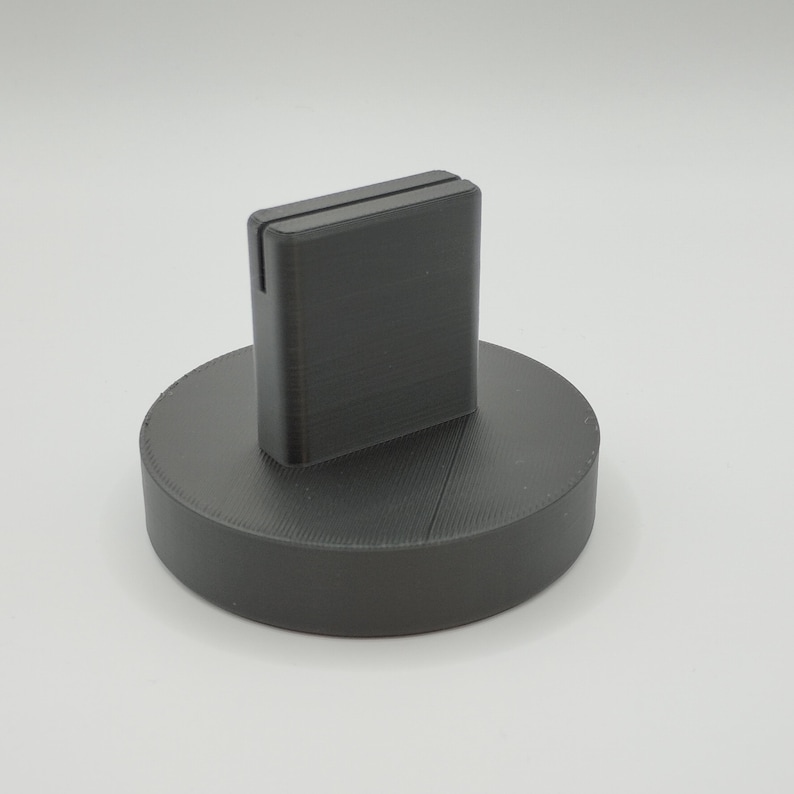 May include: A dark gray, 3D-printed phone stand. The stand has a rectangular slot to hold a phone and sits on a circular base. The stand is made of a matte material and is suitable for a desk or table.