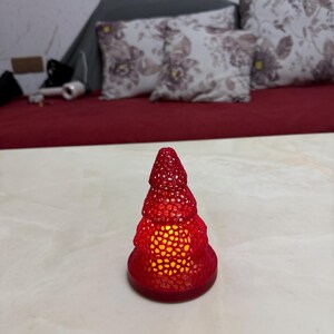 May include: A red, Christmas tree-shaped candle holder with a textured surface. The candle holder is lit from within, emitting a warm, yellow glow. The base is round and the tree shape is detailed.