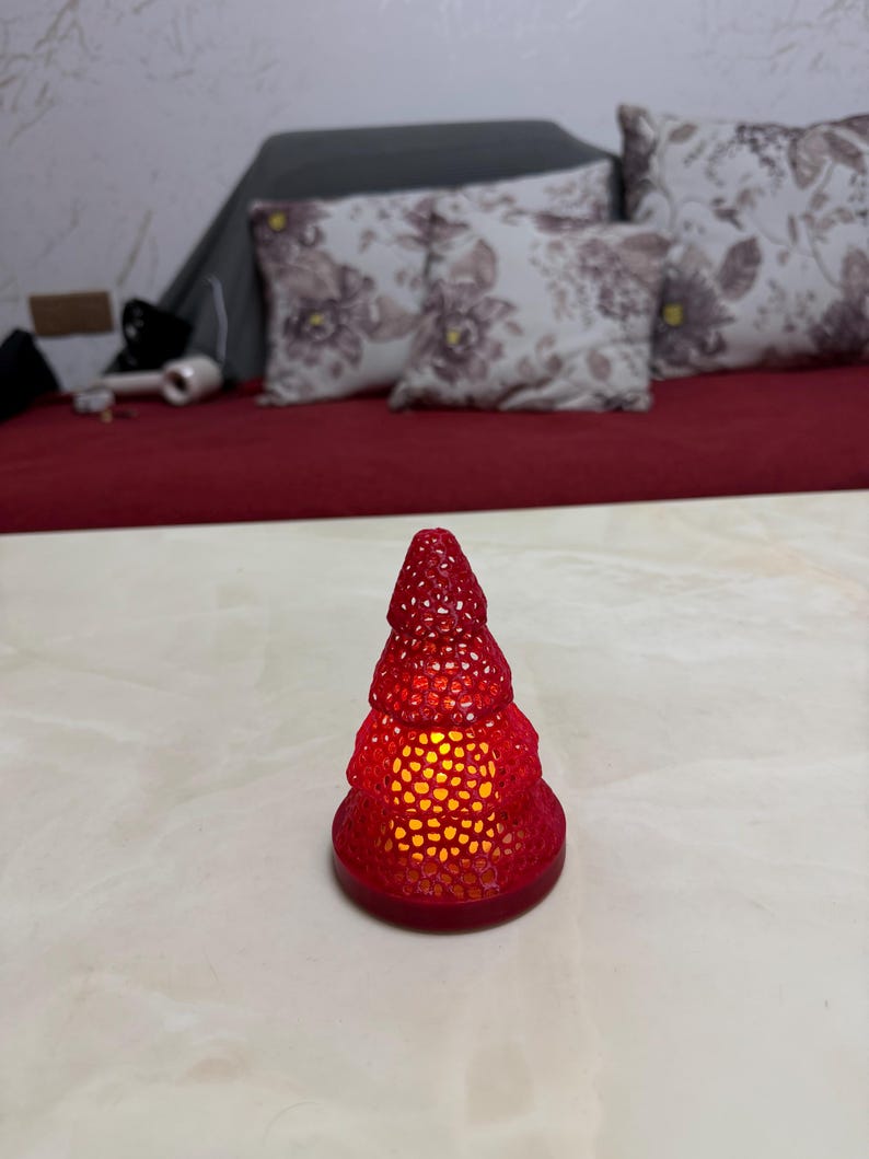 May include: A red, Christmas tree-shaped candle holder with a textured surface. The candle holder is lit from within, emitting a warm, yellow glow. The base is round and the tree shape is detailed.