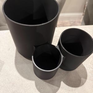 May include: Three black cylindrical containers of varying sizes. The largest is tall and slender, the smallest is short and wide, and the third is a medium size. All have a smooth, matte finish and are arranged together.