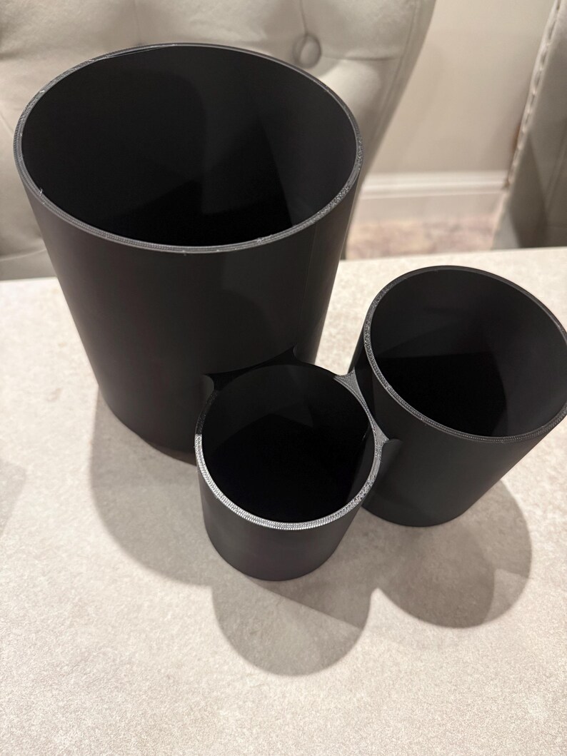 May include: Three black cylindrical containers of varying sizes. The largest is tall and slender, the smallest is short and wide, and the third is a medium size. All have a smooth, matte finish and are arranged together.