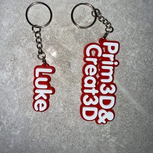 May include: Two red and white keychains. One keychain says "Luke" in white letters. The other keychain says "Prim3D & Create3D" in white letters. Both keychains have a silver key ring and chain.