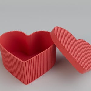 Pu&ograve; includere: Scatola a forma di cuore rosso corallo con coperchio abbinato, leggermente aperta. La scatola e il coperchio hanno una texture a coste. Lo sfondo &egrave; grigio chiaro. Ideale per regali o gioielli.