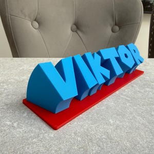 Op de afbeelding: Een blauw 3D-bord met de naam "VIKTOR" in vette, gestileerde letters. De letters zijn gemonteerd op een rode rechthoekige basis. Het bord is waarschijnlijk een decoratief item of een gepersonaliseerd cadeau.