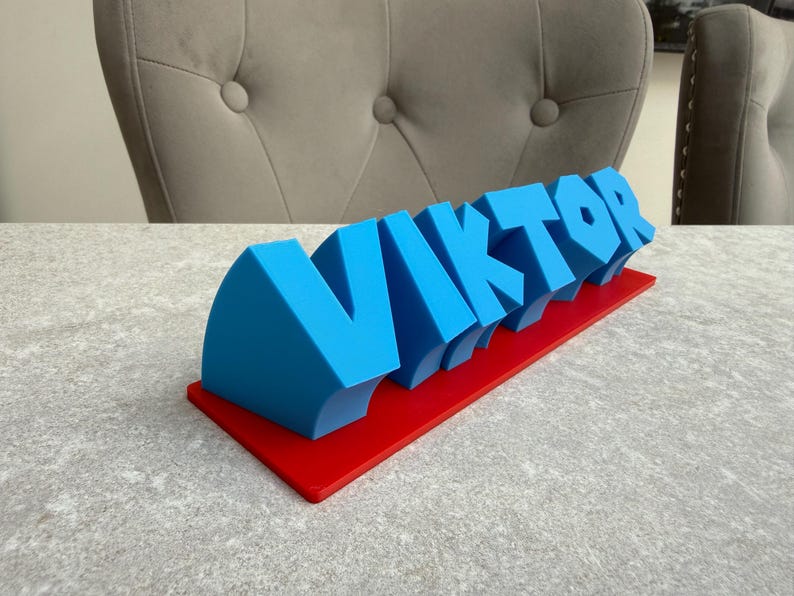 Op de afbeelding: Een blauw 3D-bord met de naam "VIKTOR" in vette, gestileerde letters. De letters zijn gemonteerd op een rode rechthoekige basis. Het bord is waarschijnlijk een decoratief item of een gepersonaliseerd cadeau.