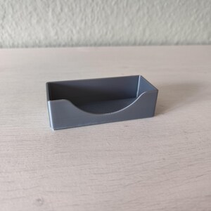 Pu&ograve; includere: Un piccolo porta biglietti da visita in plastica grigia con un bordo anteriore curvo. Il supporto &egrave; rettangolare e poggia su una superficie di legno di colore chiaro. Lo sfondo &egrave; un muro bianco sporco.