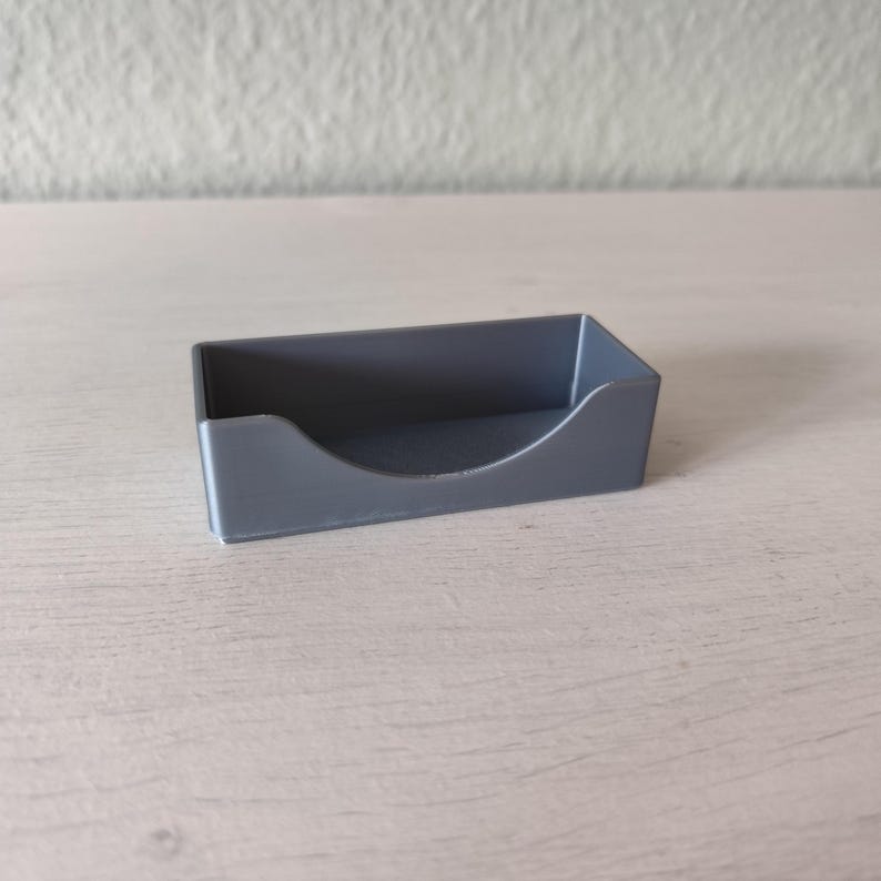 Pu&ograve; includere: Un piccolo porta biglietti da visita in plastica grigia con un bordo anteriore curvo. Il supporto &egrave; rettangolare e poggia su una superficie di legno di colore chiaro. Lo sfondo &egrave; un muro bianco sporco.