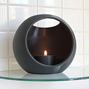 May include: A dark grey, spherical candle holder with a circular opening at the top. A lit black tea light candle sits inside, casting a warm glow. The holder is on a glass shelf against a white tiled wall.