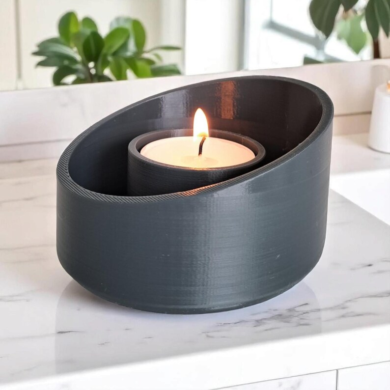 May include: A dark gray candle holder with a lit white candle inside. The holder has a unique, oval-shaped design with a textured surface. The flame is bright against the dark background, creating a warm ambiance. Home decor item.